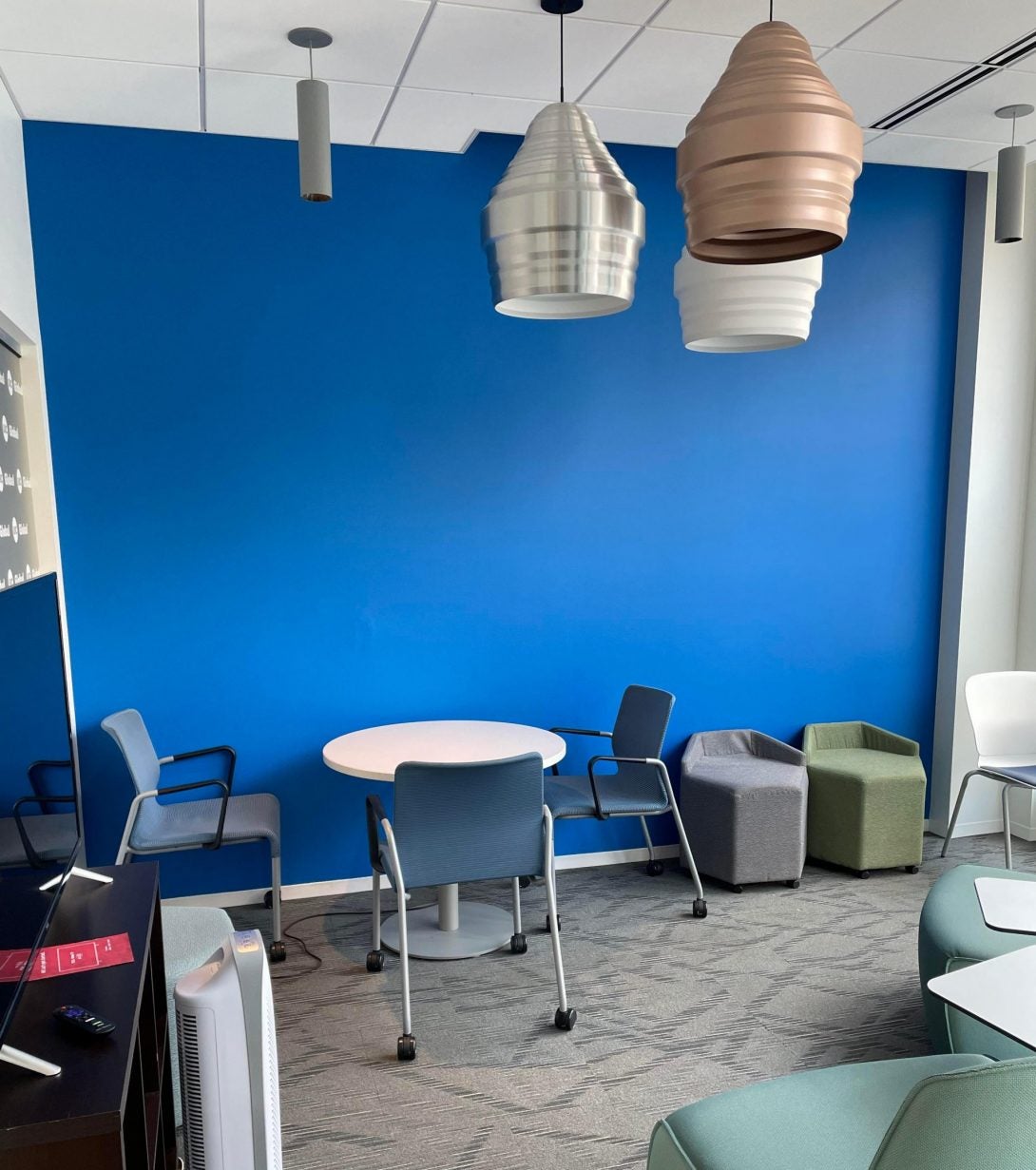 Gallery space of a blue wall with desks and hexagonal cloth chairs in front and a TV set to the left